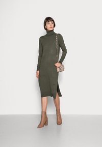 Robe turtleneck à manches longues de couleur vert olive, avec une fente latérale et un ourlet côtelé, associée à des bottes à chevilles beige et un sac à main à motifs.
