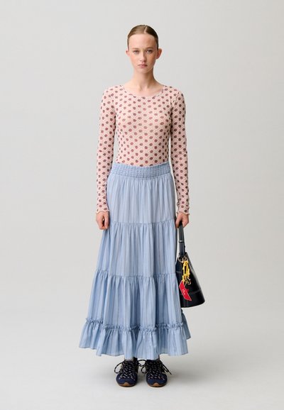 Donna con indosso un top rosa a fiori a maniche lunghe, gonna maxi azzurro chiaro a strati, sneakers blu navy, e che tiene una borsa nera con portachiavi.