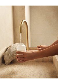 Witte cilindrische fles wordt vastgehouden onder een gouden kraan, met stromend water. Beige stenen gootsteen en een gestructureerd doek in de buurt op een glad oppervlak.