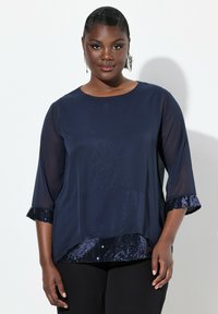 Marineblaue Bluse mit Dreiviertelärmeln, die einen glatten oberen Stoff und einen eingearbeiteten Paillettenbund aufweist. Strukturelle Akzente an den Ärmelsäumen.