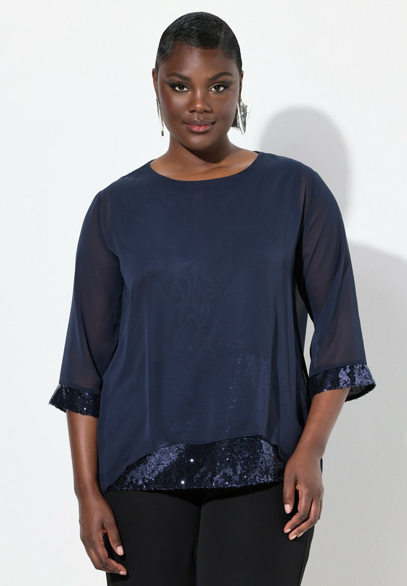 Marineblaue Bluse mit Dreiviertelärmeln, die einen glatten oberen Stoff und einen eingearbeiteten Paillettenbund aufweist. Strukturelle Akzente an den Ärmelsäumen.