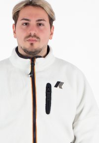 Biała polarowa kurtka z wysokim kołnierzem, zamkiem z przodu i pomarańczowym akcentem. Posiada czarną kieszeń zapinaną na zamek i logo po lewej stronie.