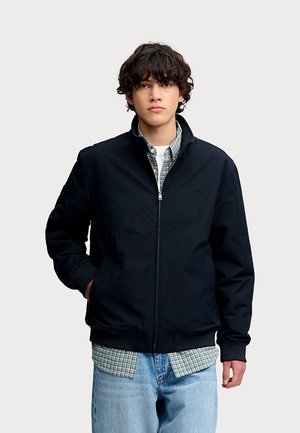 Ung mand med krøllet hår iført en sort lynlåstrøje over en grøn ternet skjorte og lyseblå jeans, stående med den ene hånd i lommen.