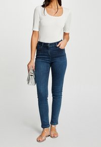 Biała ribbed top z falbankowym dekoltem i krótkimi rękawami, zestawiona z ciemnoniebieskimi skinny jeans z przednim zdobieniem w postaci ćwieków i srebrnymi sandałami.