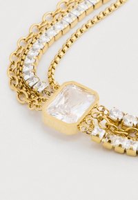 Arany lánc karkötő, amely több szálból áll, nagy téglalap alakú, tiszta drágakővel és kisebb négyzet alakú, valamint kerek tiszta kristályokkal díszítve.