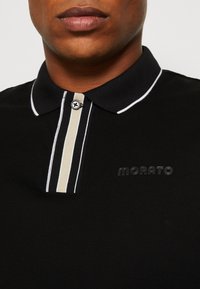 Gros plan d'une personne portant un polo noir avec col bordé blanc, bouton, et une patte verticale à rayures beige et noire, logo "MORATO".