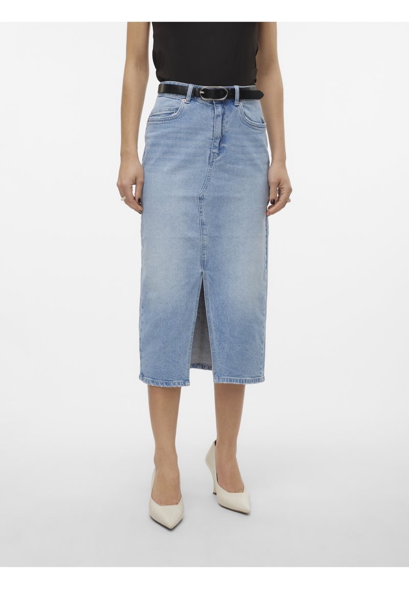 Femme portant une jupe midi en denim bleu clair avec une fente sur le devant, ceinture noire, top noir et talons blancs pointus, debout devant un fond blanc.