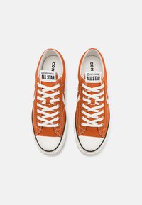 Oranžs kanvas sporta apavi ar baltiem šņorēm, melnu gumijas zoli un klasisko "All Star" zīmolu uz mēles. Pievienots balts zvaigznes logo.