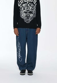 Czarny sweter z grafiką tygrysa, w połączeniu z niebieskimi szerokimi spodniami dżinsowymi z białym napisem "Ed Hardy" na lewej nodze i czarnymi butami.