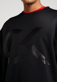 Sweatshirt noir avec un logo en caoutchouc noir proéminent, col rond et finition texturée. Superposé avec une chemise rouge en dessous.