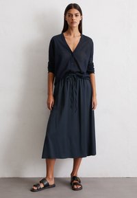Marineblaue Strickjacke mit V-Ausschnitt und Knöpfen, kombiniert mit einem fließenden Maxikleid mit elastischem Bund und Kordelzug. Flache schwarze Sandalen.