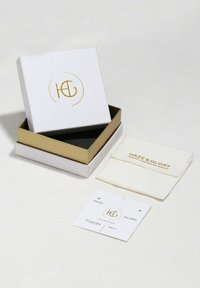 Boîte blanche avec des accents dorés, comportant le logo "HG". Comprend un plateau intérieur noir, un sac à bijoux et une carte d'entretien. Texture lisse, design minimaliste.