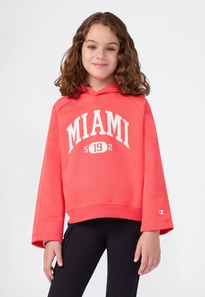 Niña joven con pelo rizado que lleva una sudadera color coral con el texto "MIAMI 192" y pantalones negros, de pie contra un fondo blanco liso.