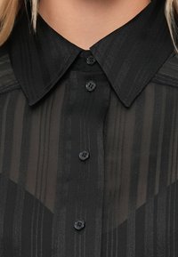 Svart skjorte med knappestolpe, med en teksturert stripedesign, semi-transparent stoff, spiss krage og svarte knapper.