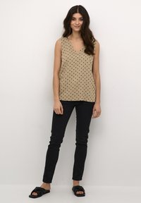 Haut sans manches beige avec des pois noir, design en V, associé à un jean skinny noir et des sandales glissantes noires. Texture de tissu lisse.
