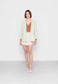 Blazer verde claro sobre uma blusa castanha, combinado com uma saia floral rosa. Modelo usa saltos brancos com tiras. Fundo minimalista.