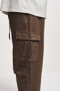 Pantaloni cargo in tessuto marrone scuro, con una grande tasca laterale e una finitura testurizzata. Abbinati a una camicia bianca, che mostra un design a strati.