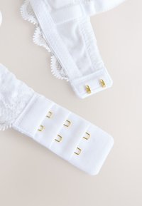 Spallina di reggiseno in pizzo bianco con chiusure con gancio e occhiello color oro, mostrata slacciata su sfondo beige chiaro.