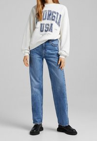 Pull blanc avec un motif "Georgia USA" ; jean bleu taille haute  à coupe droite ; bottes noires épaisses ; look lisse et décontracté.