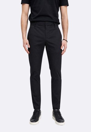 Uomo che indossa pantaloni neri slim fit, t-shirt nera, sneakers nere con suola bianca e un braccialetto nero al polso destro, in piedi dritto.