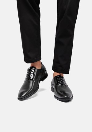 Scarpe nere eleganti con una finitura texture e a motivo, caratterizzate da accenti lucidi e lacci tradizionali, abbinate a pantaloni neri con risvolto.