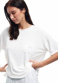 Jonge vrouw met lang donker haar kijkt naar beneden terwijl ze een losse witte t-shirt draagt met een kleine bloemenzak en lichtpatronenbroek.