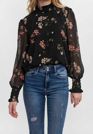 Blouse noire transparente avec imprimé floral dans des nuances de rose et de vert. Longues manches bouffantes avec des poignets froncés, col haut à volants. Porté avec un jean bleu.