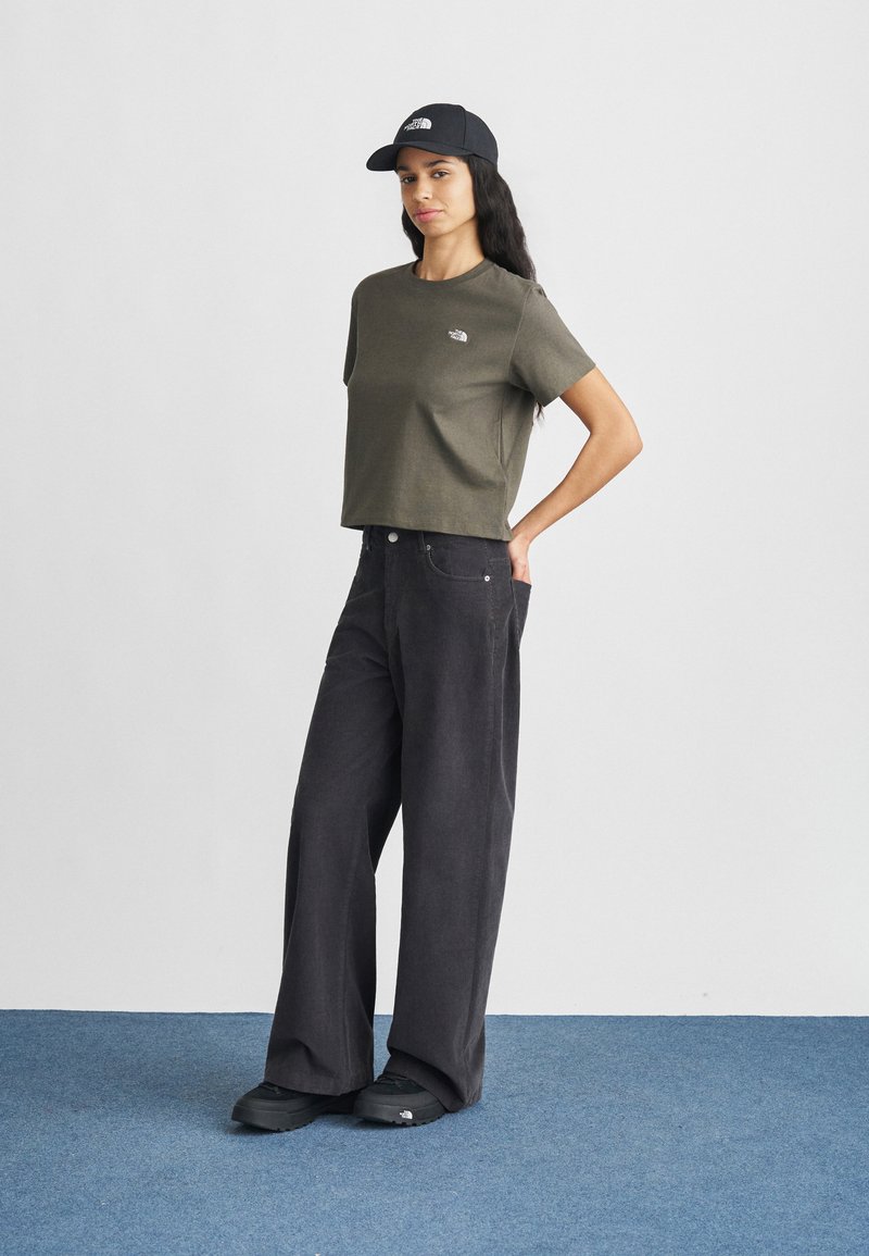 Dark grey wide-leg pants, olive green cropped t-shirt with a logo, and black cap with logo. Person stands on blue carpeted floor.