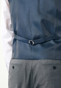 Vista posteriore di una persona che indossa un gilet blu navy a fantasia con cinturino regolabile sopra una camicia bianca e pantaloni grigi con tasche con bottoni.