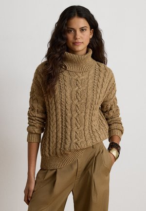 Femme aux longs cheveux foncés bouclés portant un pull à col roulé en maille torsadée marron et un pantalon marron taille haute, avec des bracelets au poignet.