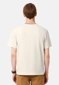 T-shirt à manches courtes de couleur crème avec un col rond, comportant un accent rayé marron et noir au niveau du cou et une coupe décontractée.