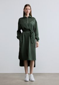 Πράσινο φόρεμα με μακριά μανίκια και φερμουάρ, με ζώνη στη μέση και δύο τσέπες στο στήθος· κατασκευασμένο από λείο ύφασμα· συνδυασμένο με λευκά αθλητικά παπούτσια.