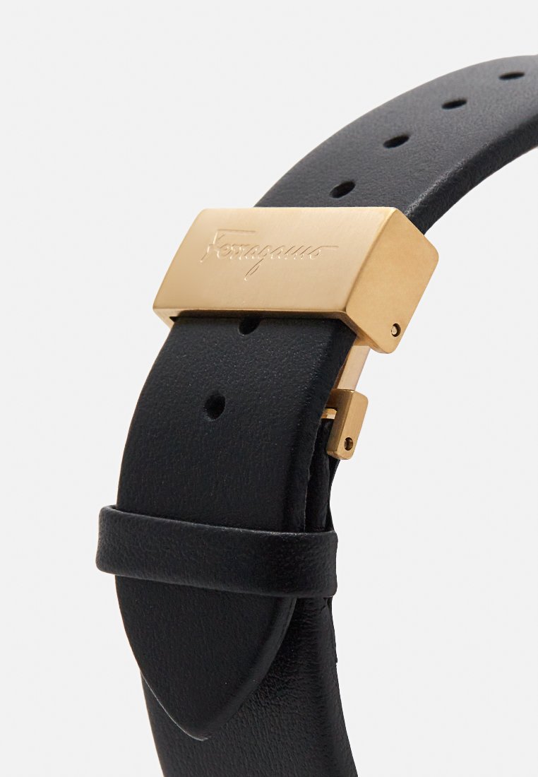 Bracelet de montre en cuir noir avec une boucle en métal doré gravée du logo. Le bracelet dispose de plusieurs trous d'ajustement pour la taille.