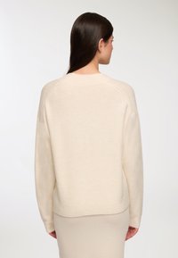 Beżowy sweter wełniany z okrągłym dekoltem, luźnym krojem i ściągaczami na mankietach. Gładka struktura i minimalne detale przeszycia na ramionach.