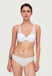 Mulher em pé, de pé, usando um sutiã branco com detalhes em renda e uma lingerie branca estilo biquíni, mostrando uma tatuagem de rosa no antebraço esquerdo.