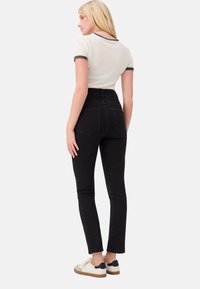 Donna bionda che indossa una maglietta bianca a maniche corte con rifiniture nere, jeans neri a vita alta e sneaker bianche, rivolta di spalle su uno sfondo bianco.