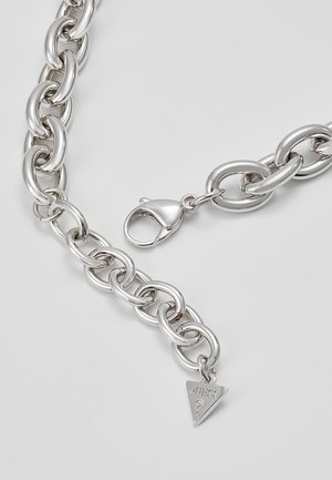 Silbernes Kettenarmband mit großen ovalen Gliedern und einem Karabinerverschluss, versehen mit einem dreieckigen Guess-Logo-Anhänger vor hellgrauem Hintergrund.