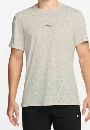 Homme portant un t-shirt beige à manches courtes Nike Running Division avec un imprimé léopard discret et un short noir Nike sur un fond uni.