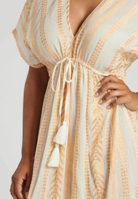 Robe en coton avec un col en V et une taille à nouer, ornée de motifs géométriques orange et d'accents en franges blanches. Tissu léger et fluide.