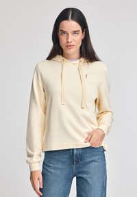 Cremefarvet hættejakke med snor, ribbet manchetter og et lille logo på brystet. Sammen med blå denimjeans. Blød tekstur.