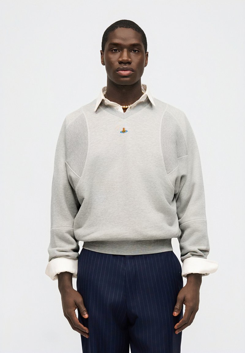 Giovane uomo che indossa un maglione grigio chiaro sopra una camicia bianca con collo e pantaloni blu scuro a righe sottili, in piedi davanti a uno sfondo bianco uniforme.