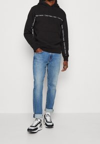 Sudadera negra con el logo blanco de "Calvin Klein Jeans" en el pecho y las mangas, combinada con jeans de color azul claro y zapatillas negras y blancas.