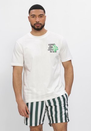 Mann trägt ein weißes T-Shirt mit grünem Text und Design, kombiniert mit grün-weißen, vertikal gestreiften Shorts, steht vor einem einfarbigen Hintergrund.