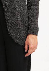 Šedý pletený cardigan s texturovaným materiálem, otevřeným předním dílem a žebrovanými manžetami, nošený na černém spodním oblečení.