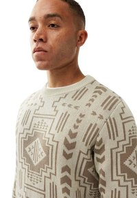 Mann mit kurzen Haaren und Ohrring, der einen beigen Pullover mit geometrischen Tribal-Mustern trägt und nach vorne blickt, vor einem weißen Hintergrund.