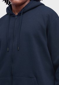 Ciemnogranatowa bluza z kapturem na zamek błyskawiczny z troczkami, noszona przez osobę o kręconych włosach, pokazująca górną część tułowia i część twarzy.