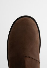 Stivale in suede marrone con punta tondeggiante. Texture liscia, cuciture nere e suola sottile e leggermente rialzata. Design minimalista, senza hardware visibile.