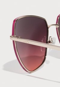 Sunglasses with a large, angular shape. Dark gradient lenses, gold metal frame, and pink accents along the top edge. Engraved logo on hinge.