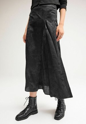 Femme portant une jupe midi noire texturée associée à des bottines noires à lacets et un pull côtelé noir.