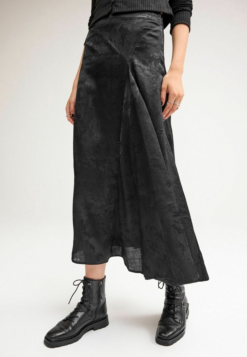 Femme portant une jupe midi noire texturée associée à des bottines noires à lacets et un pull côtelé noir.